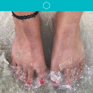 Woman's feet on sands with wave splashing over toes.  She's wearing a transform mindfulness temporary tattoo on her foot.